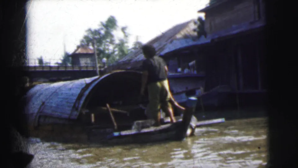 Calcutta home-movie frame of an adult on a narrow boat near a larger covered boat and water-edge buildings.