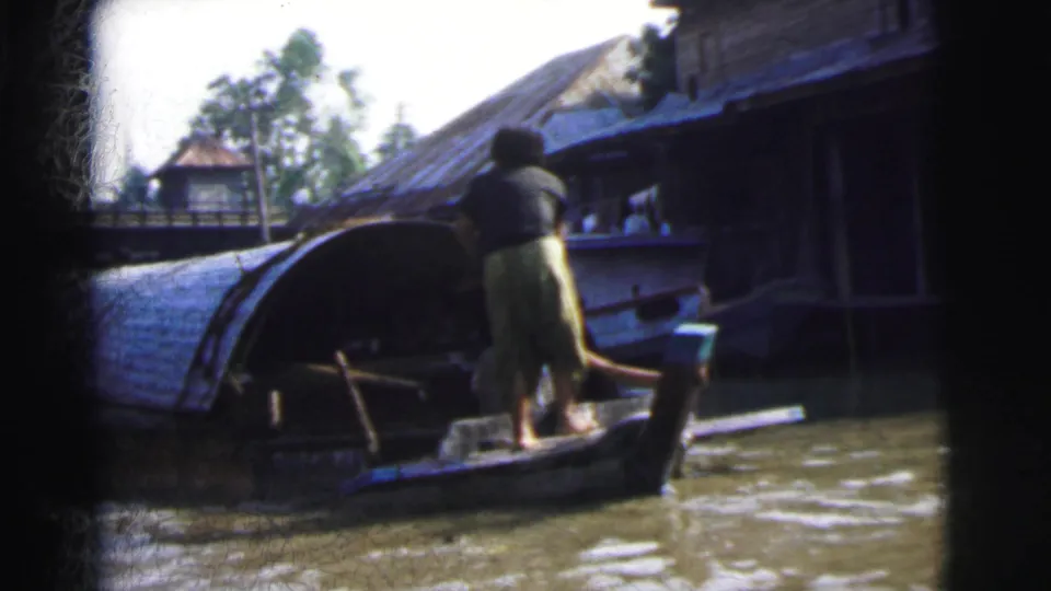 1962 Calcutta 8mm frame showing an adult steering or standing on a small boat near covered craft and stilt-like waterfront buildings.