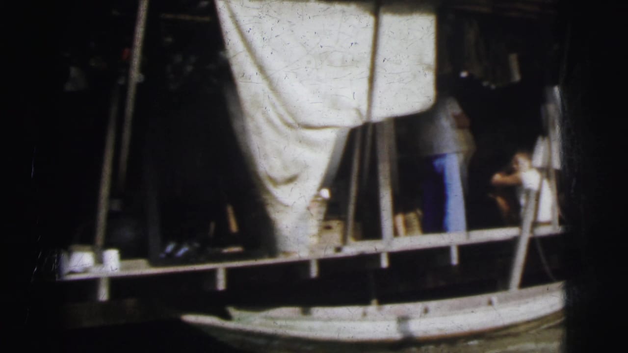 1962 8mm home-movie frame showing people on a shaded river platform with a boat hull, poles, and fabric or roof cover nearby.