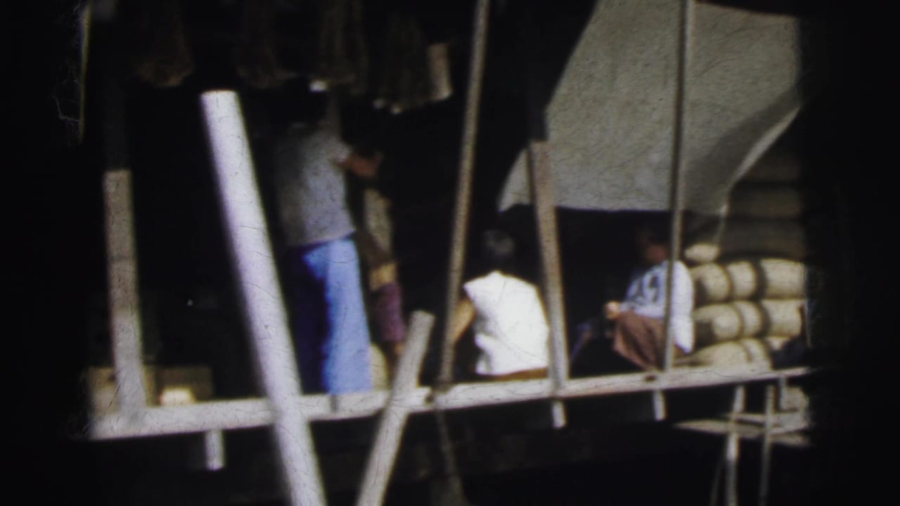 1962 archive frame of people gathered around a waterside wooden platform with poles, boat structure, and stacked cylindrical forms at right.