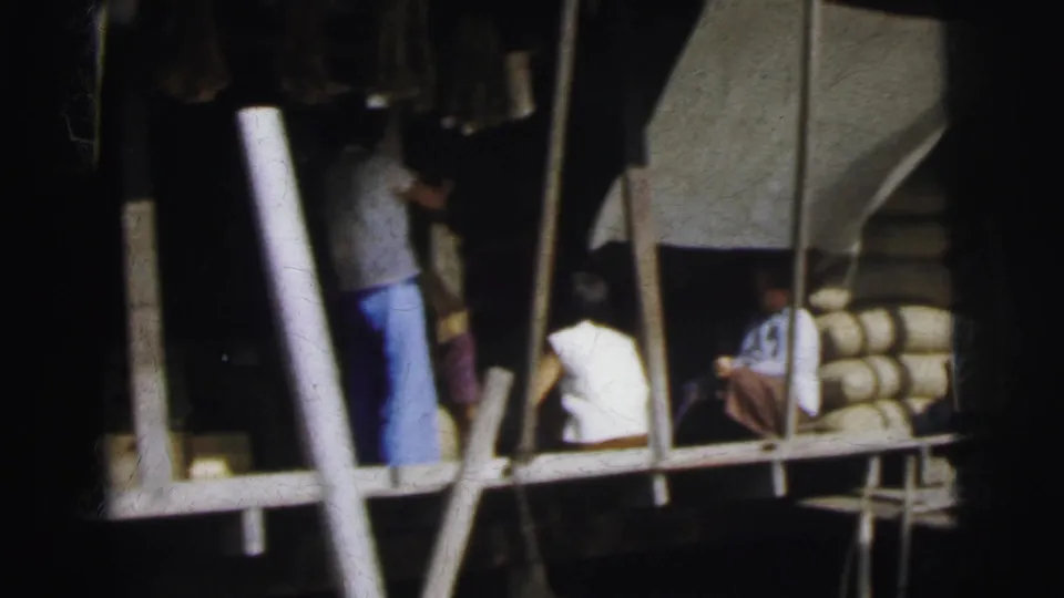 1962 archive frame of people gathered around a waterside wooden platform with poles, boat structure, and stacked cylindrical forms at right.