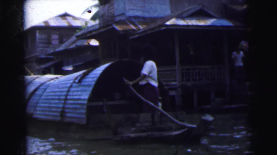 1962 home-movie view of a person propelling a small boat by pole near corrugated roofing and wooden riverside houses.