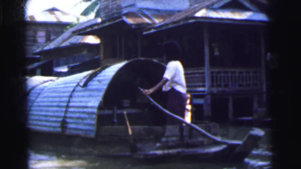 1962 8mm frame showing a person guiding a small boat by pole in front of wooden waterside houses with corrugated roofs.