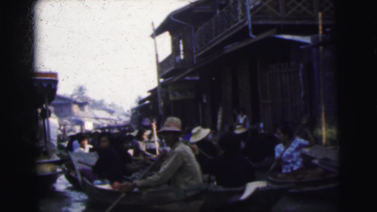 Crowded boats pass near stilt-like wooden buildings along a canal.