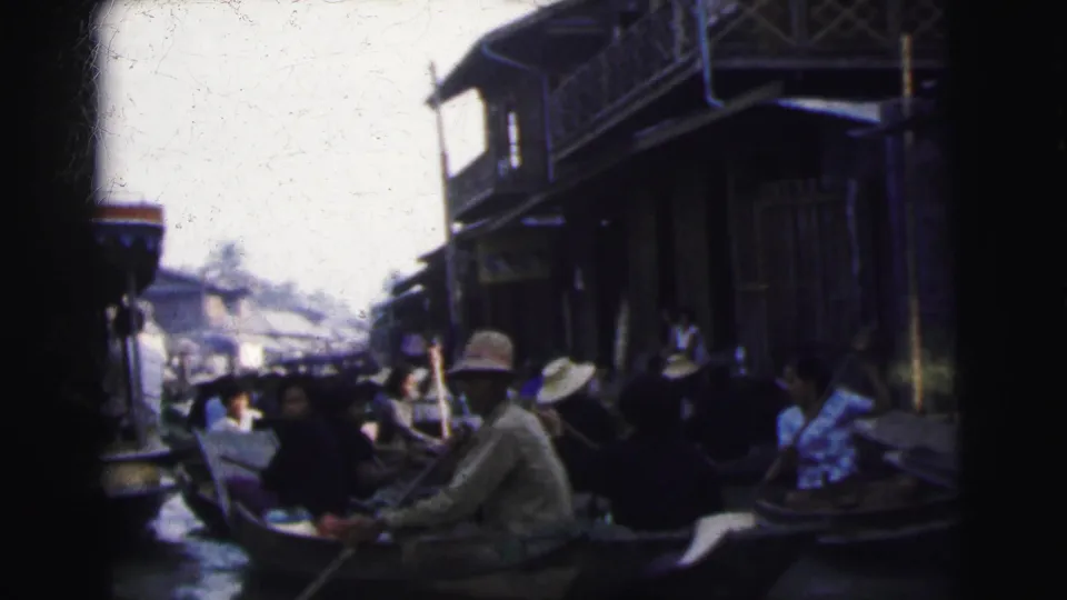 1962 Calcutta footage frame with several people seated in boats near canal-side wooden structures and a paddler in a hat.