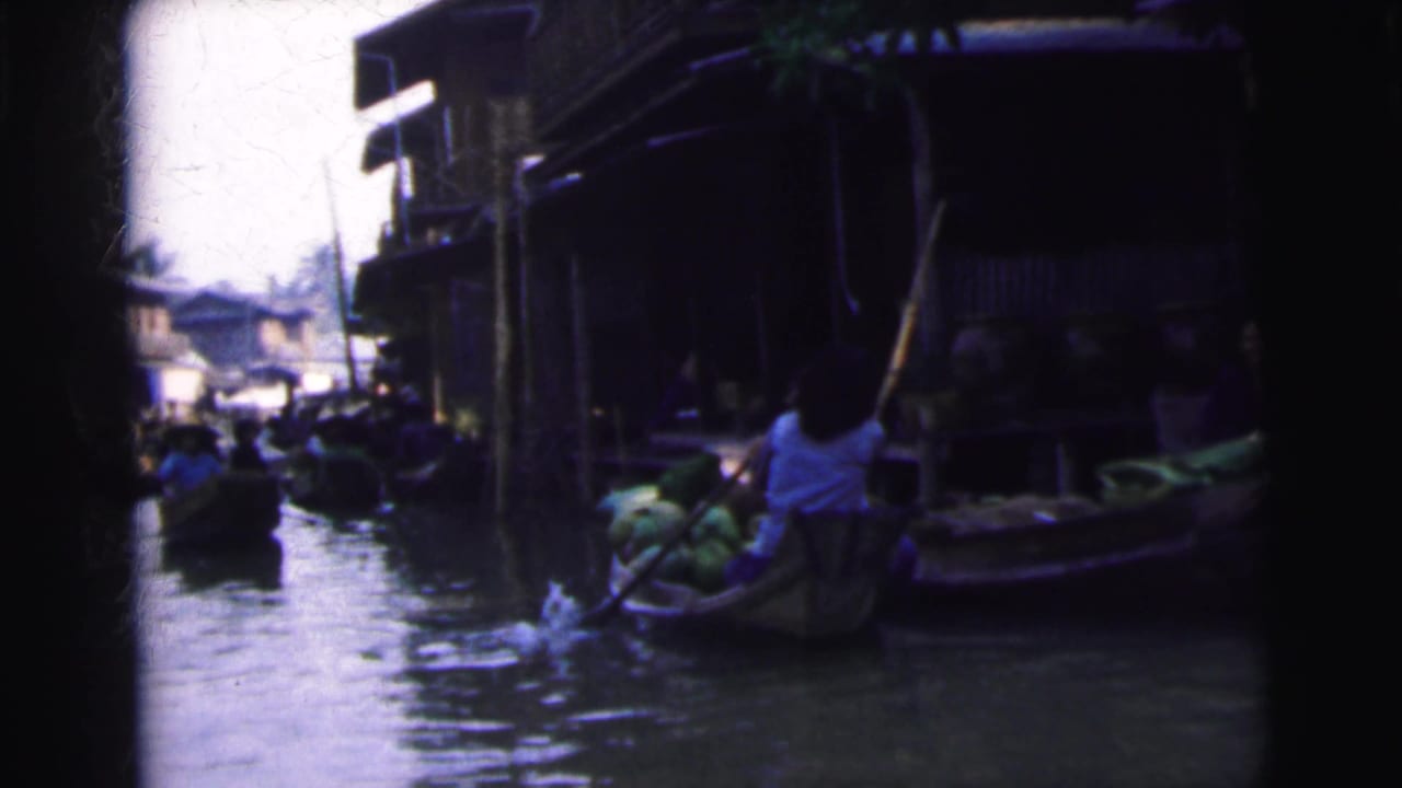Wooden boats move through a narrow waterway beside riverside buildings.