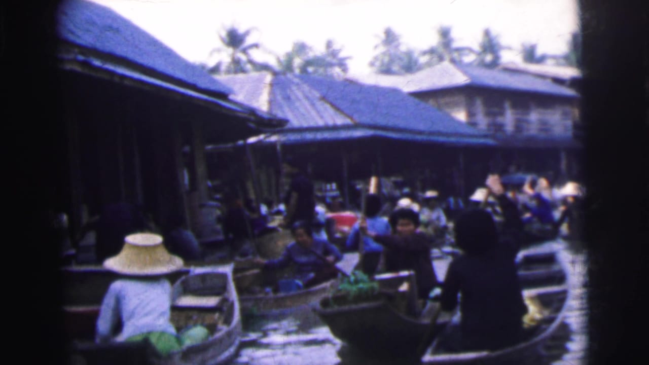 Boats pass through a narrow market waterway beside buildings and palm trees.