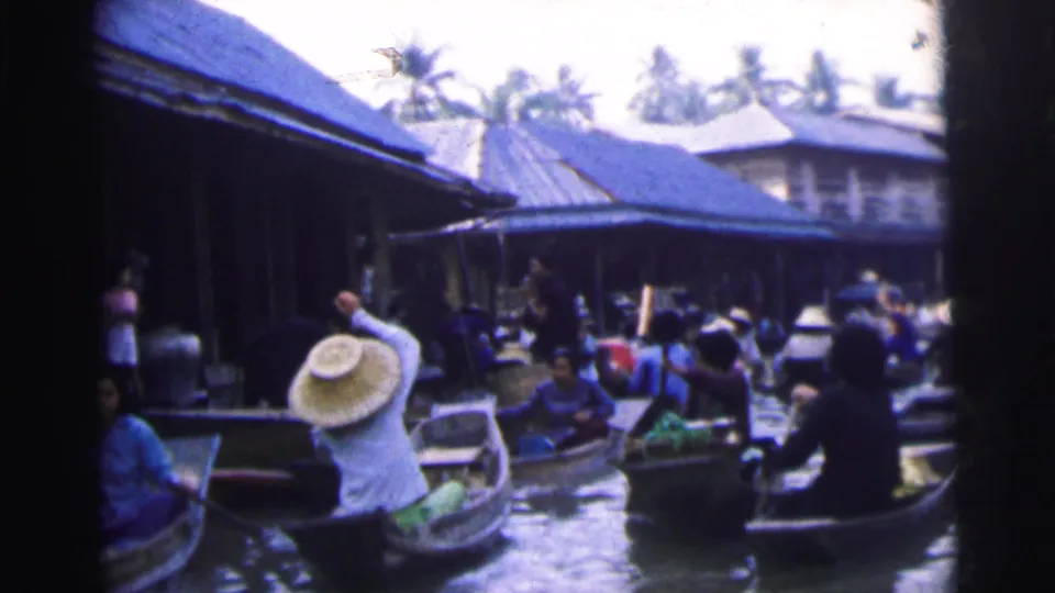 1962 8mm frame from Calcutta showing a person in a conical hat rowing near other boats in a busy river market.