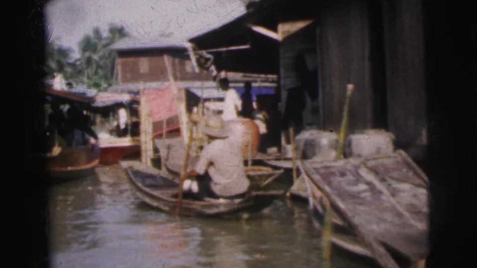 1962 Calcutta canal view with a seated adult paddling a small wooden boat past stilt-like buildings and moored craft.
