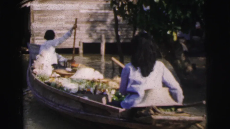 A produce boat on a Calcutta canal carries leafy vegetables and goods while two people sit and row near wooden waterside buildings.