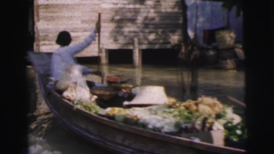 A person in light clothing rows or poles a produce-laden boat along the Calcutta canal, with a wooden building raised above the water behind.