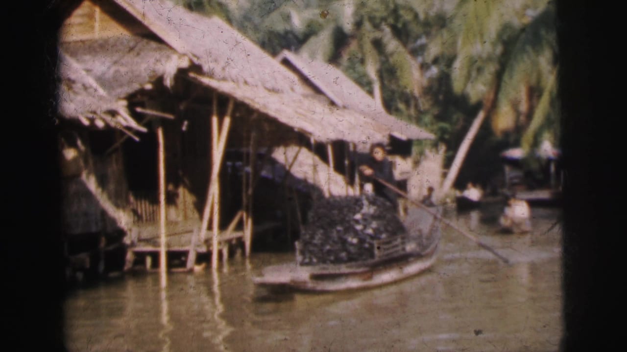 1962 8mm home-movie frame showing thatched waterside homes, palm trees, and a narrow boat carrying stacked goods along a river or canal.