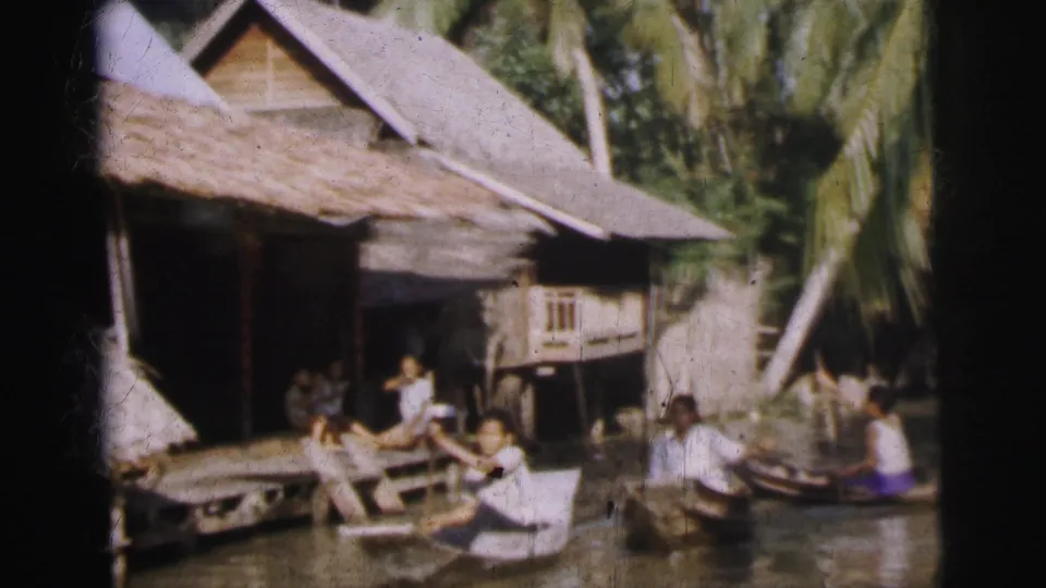Home-movie frame showing people near small wooden boats beside thatched waterside homes, with palms and dense greenery behind them.