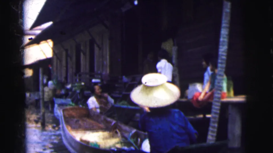 A close 8mm frame shows a person wearing a conical straw hat in a boat beside a dark wooden riverside structure, with other figures nearby.