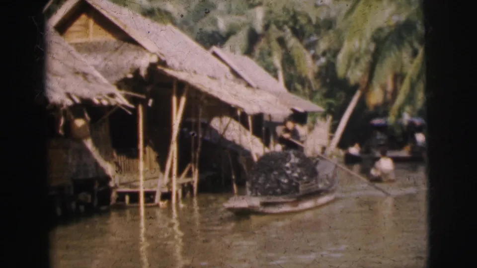 1962 Calcutta riverbank frame showing a thatched stilt house, a small canoe, water and people near the shore.