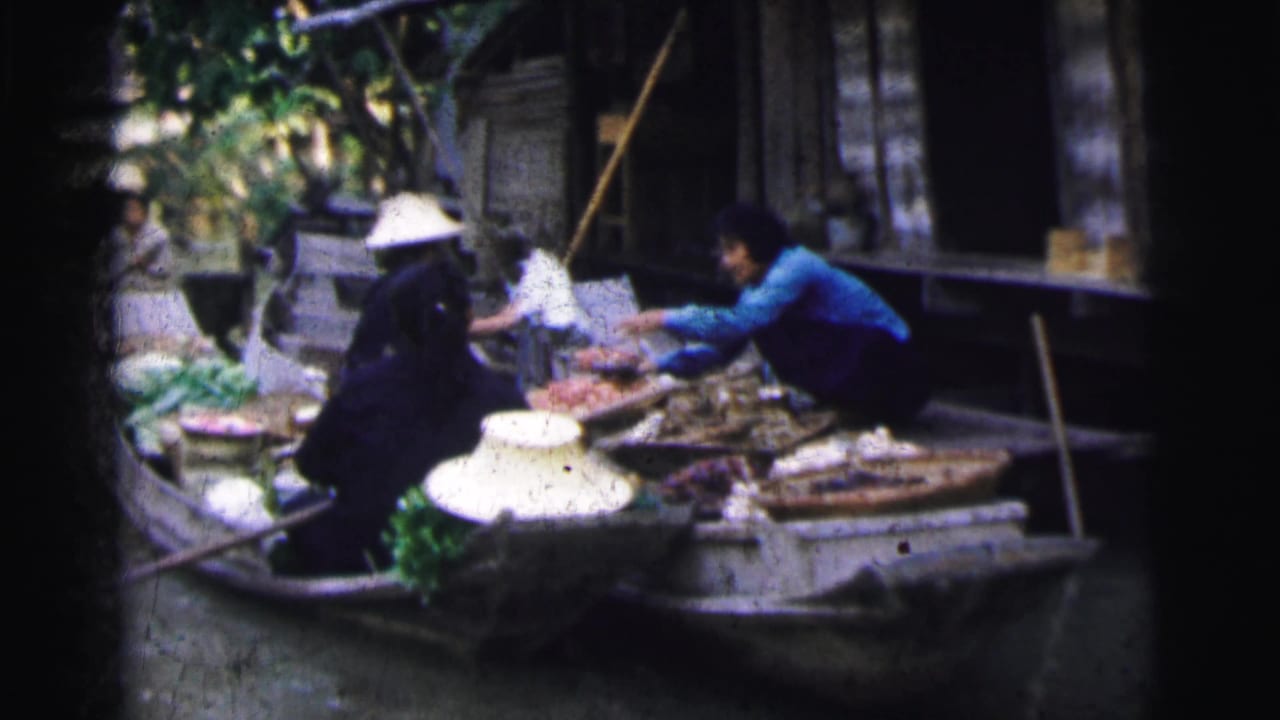 Vendors in small boats exchange goods beside a canal-side structure.