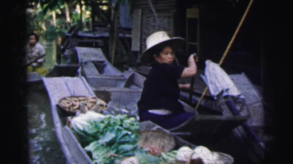 1962 Calcutta home-movie frame showing a vendor in a woven hat paddling beside boats loaded with vegetables and baskets.
