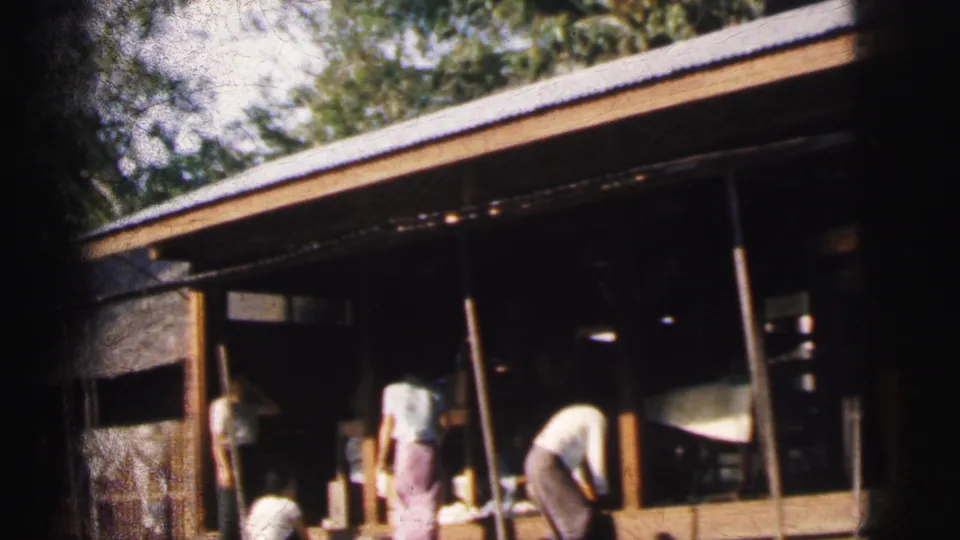 1962 Calcutta home-movie frame showing adults near a shaded wooden riverside dwelling or work area.
