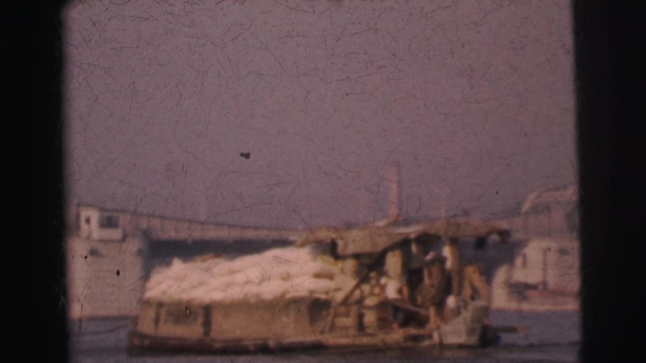 1962 Calcutta home-movie frame showing a loaded river barge with sacks and a bridge beyond the water.