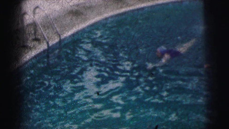 The swimmer nears the edge of the frame in 1962 Calcutta pool footage, with scratches, grain, and dark gate edges visible.