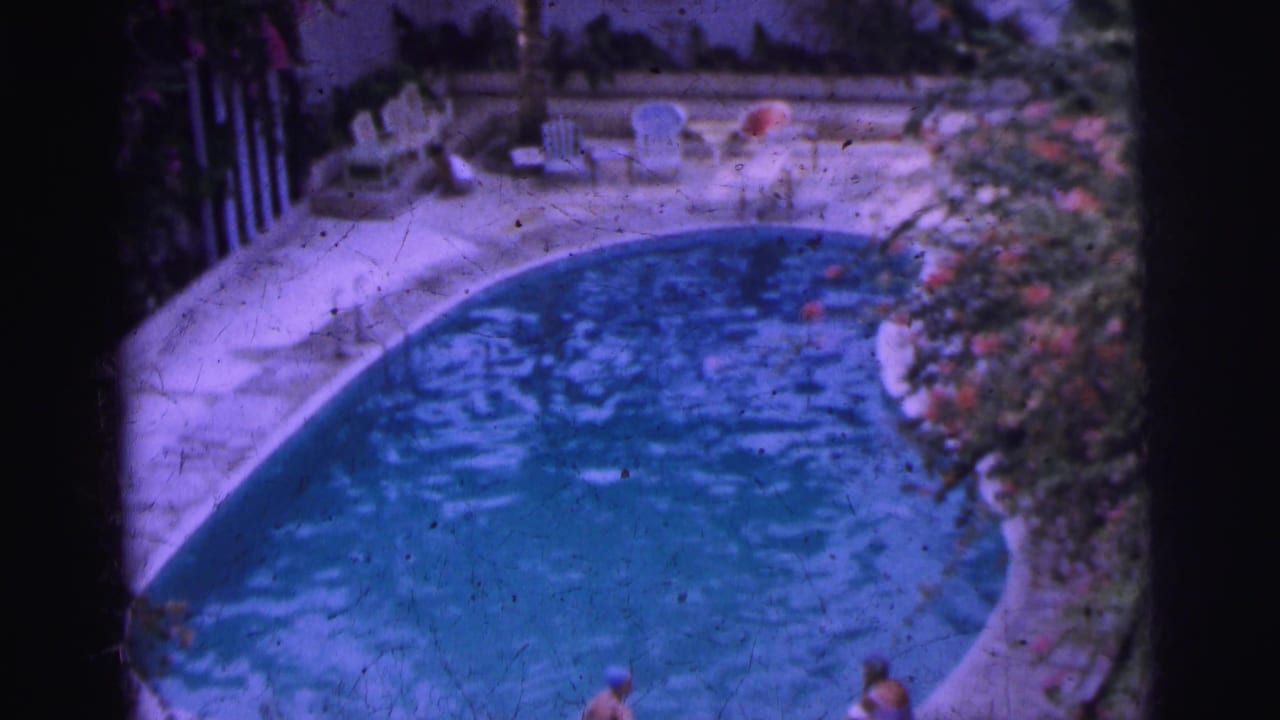 1962 Calcutta home-movie frame showing a kidney-shaped pool, white chairs, flowering shrubs, and swimmers near the edge.