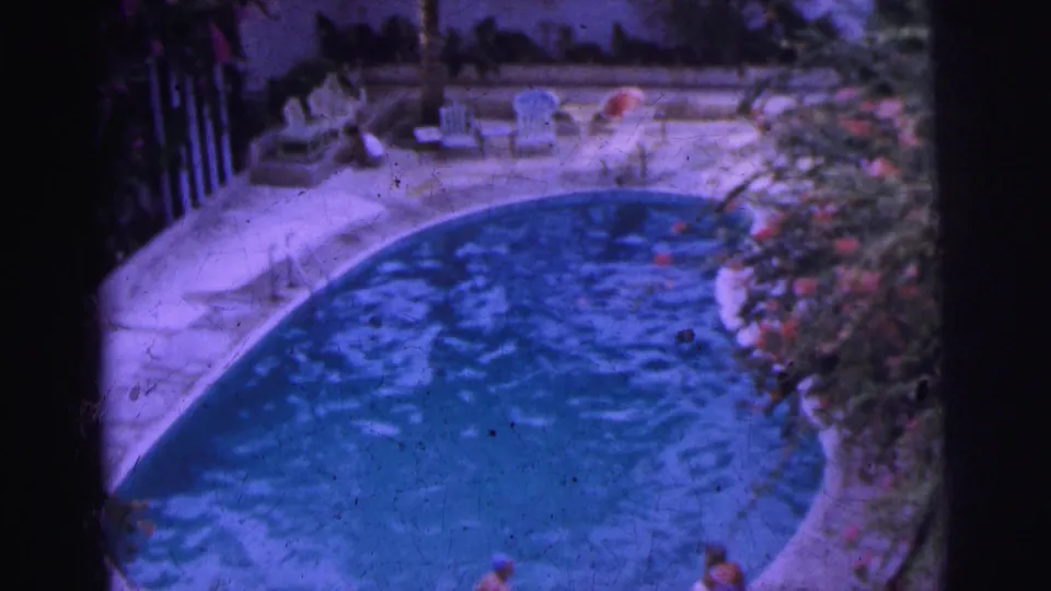 1962 Calcutta home-movie frame of a curved pool bordered by lounge chairs, plants and people partly visible near the foreground.