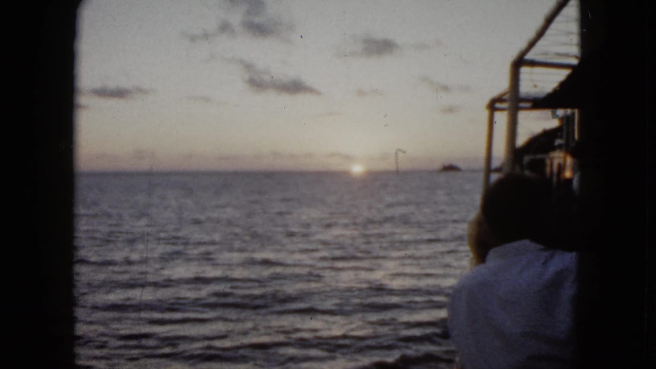 1962 Acapulco home-movie frame showing a man from behind on a boat, with sunrise light on the ocean and distant land on the horizon.