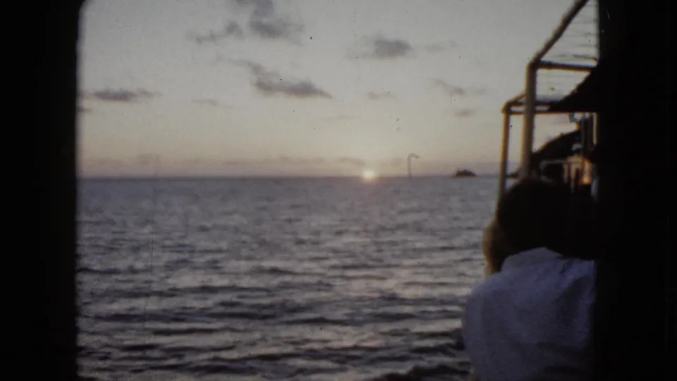 1962 Acapulco home-movie frame showing a man from behind on a boat, with sunrise light on the ocean and distant land on the horizon.