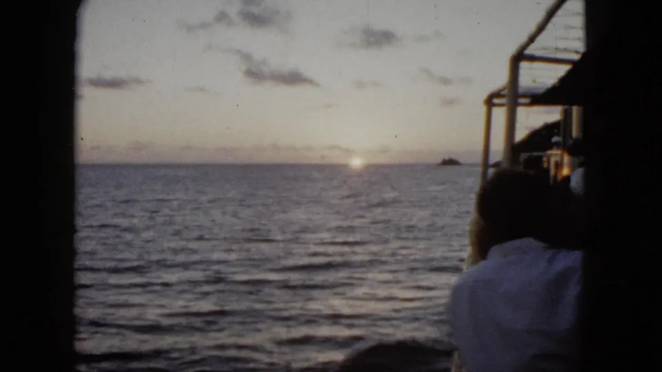1962 Acapulco frame with rippled ocean, low sun near the horizon, a distant landform, and a man partly visible at the boat edge.