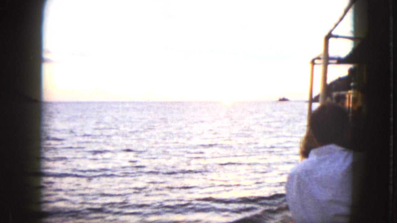 Passenger seen from behind on a boat in Acapulco as the low sun reflects on the ocean, with a distant rock on the horizon.