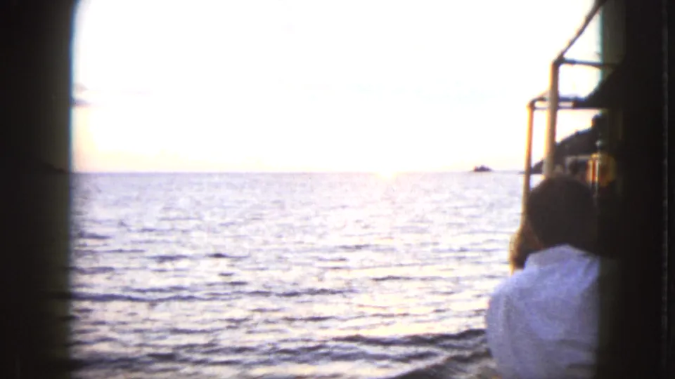 Passenger seen from behind on a boat in Acapulco as the low sun reflects on the ocean, with a distant rock on the horizon.