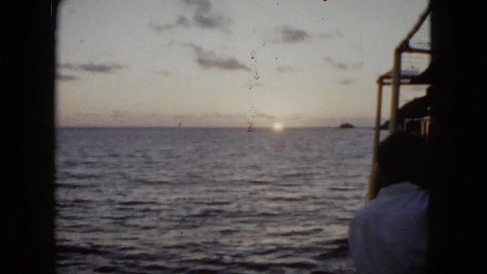 8mm view from a boat in Acapulco showing a passenger at right, calm ocean, low sun reflection, clouds, and a distant rock.