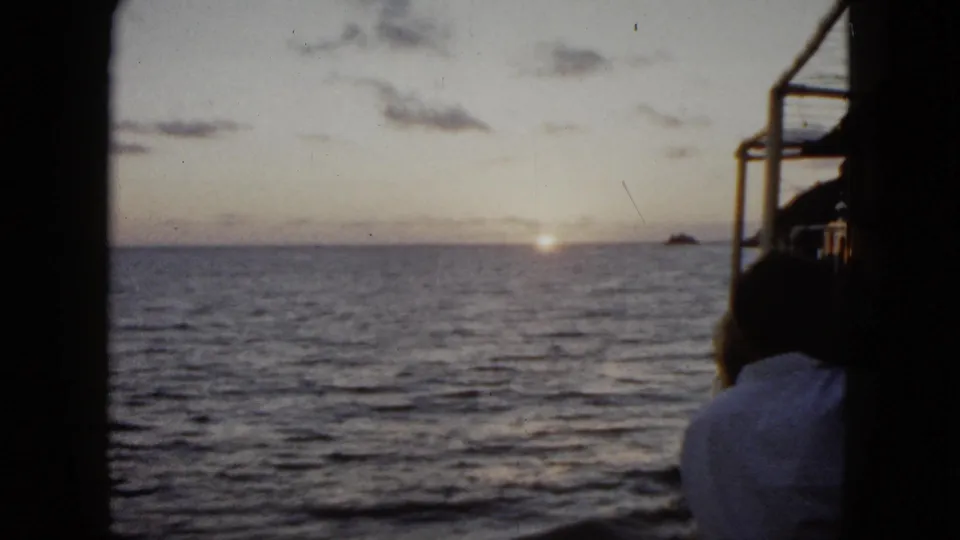 1962 Acapulco boat footage showing the sun near the horizon, rippled ocean, distant rock, and a passenger partially visible at right.