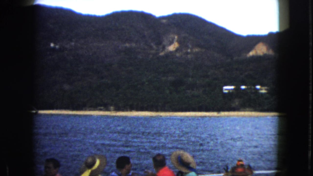 1962 Acapulco boat-tour view with passengers in hats below a blue bay, sandy shore, and forested mountain backdrop.