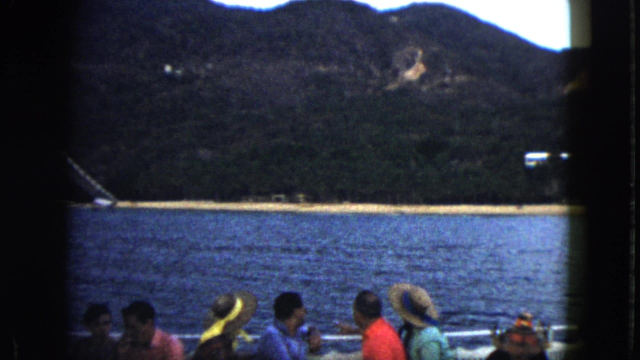 Acapulco home-movie frame showing boat passengers in summer clothing before calm water, beach, and dark hillside.
