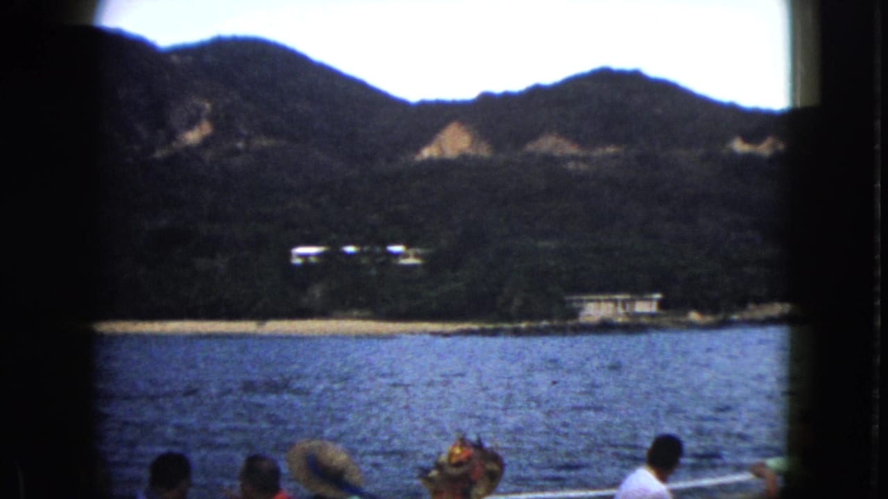 1962 Acapulco boat view with passengers at the bottom of frame, blue water, a sandy shore, white buildings, and mountain slopes beyond.