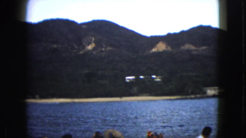 Soft 8mm frame of the Acapulco coast, showing bay water, beach, hillside vegetation, small white buildings, and boat passengers in the foreground.