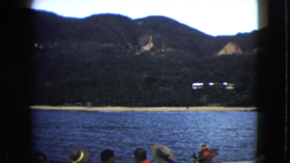 1962 Acapulco home-movie frame from a boat, with passengers in the foreground and a beach, hillside, and small white buildings across the bay.