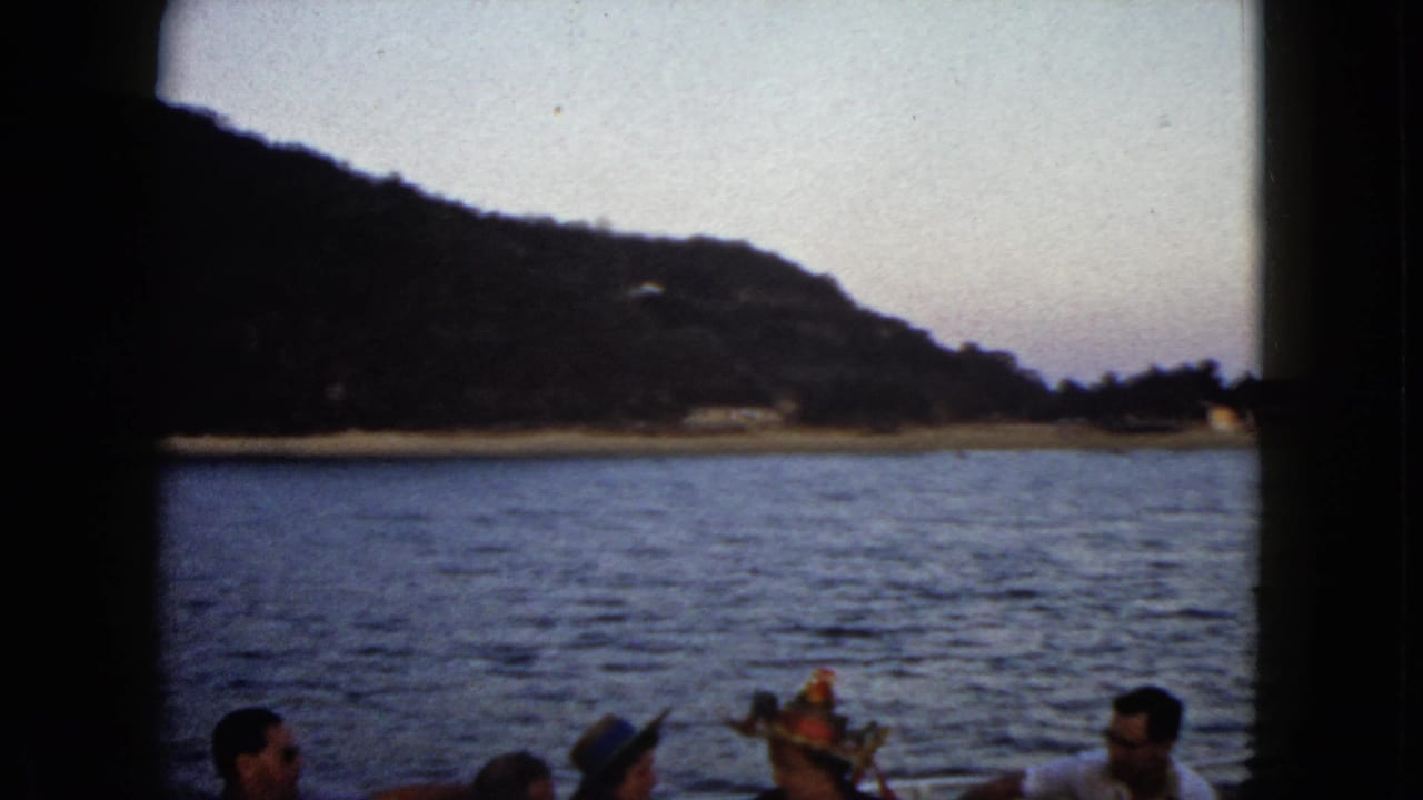 1962 Acapulco home-movie frame showing passengers on a boat with blue water, a low shoreline, and mountains in the distance.