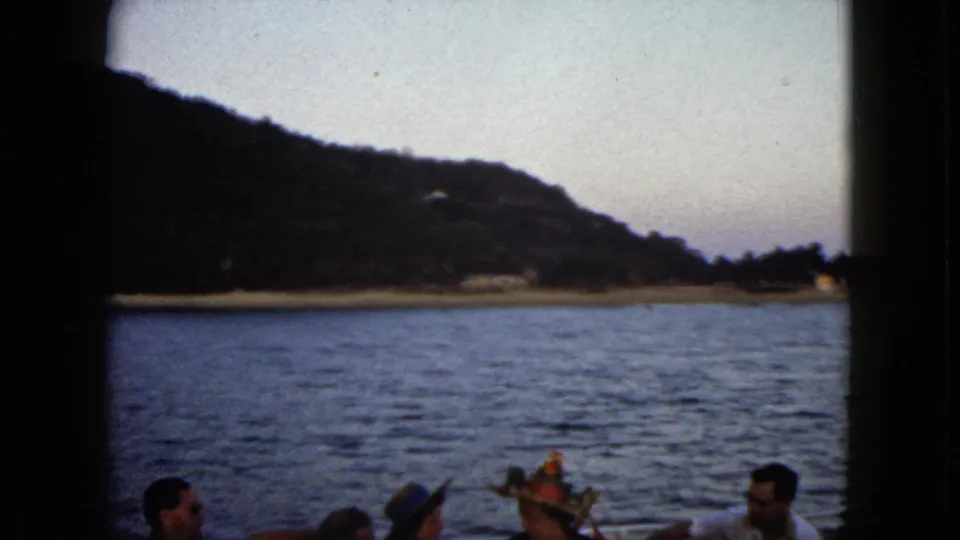 1962 Acapulco home-movie frame showing passengers on a boat with blue water, a low shoreline, and mountains in the distance.