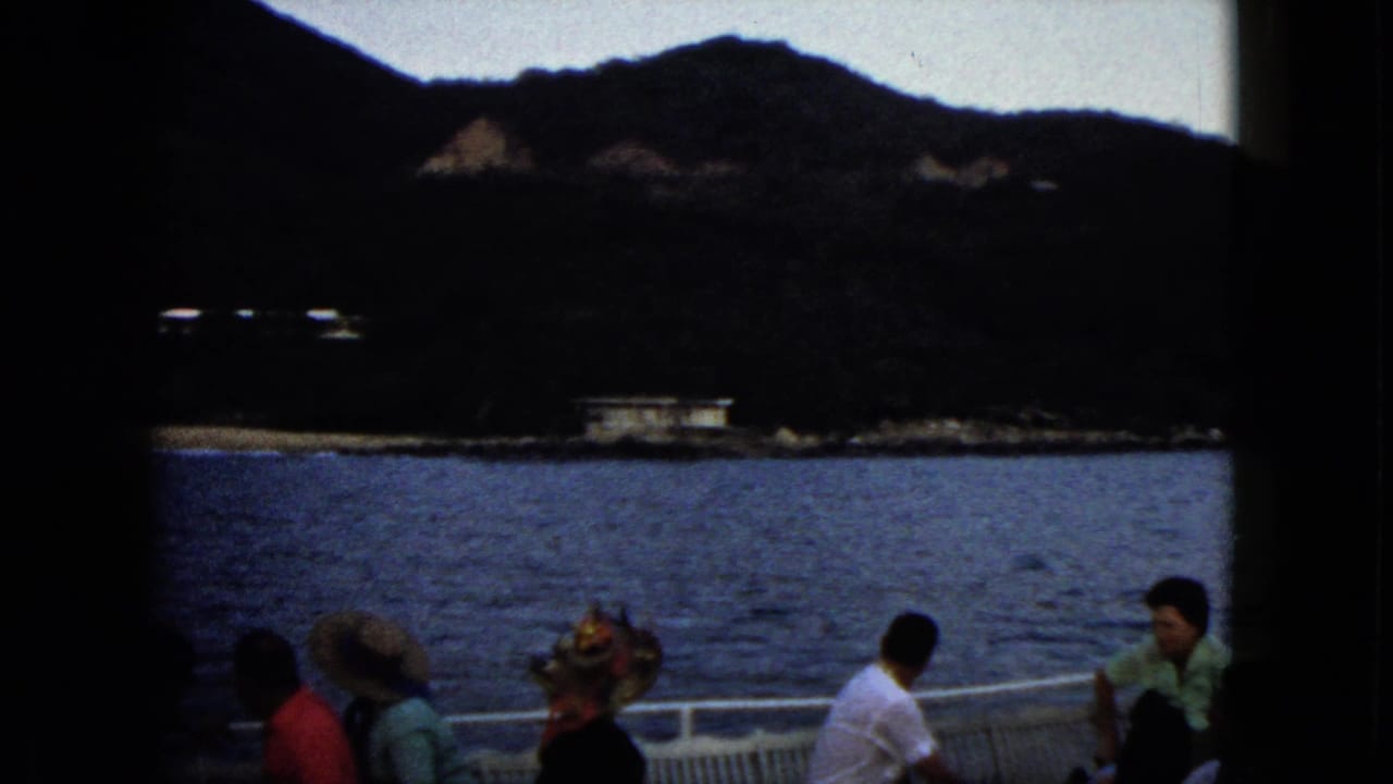Passengers seated beside a boat rail with blue water, shoreline buildings, and mountains behind them.