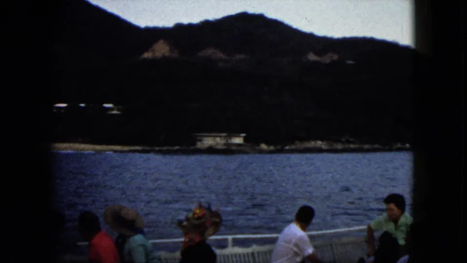1962 Acapulco boat-tour frame with seated passengers in hats and summer clothing against coastal water and wooded mountains.