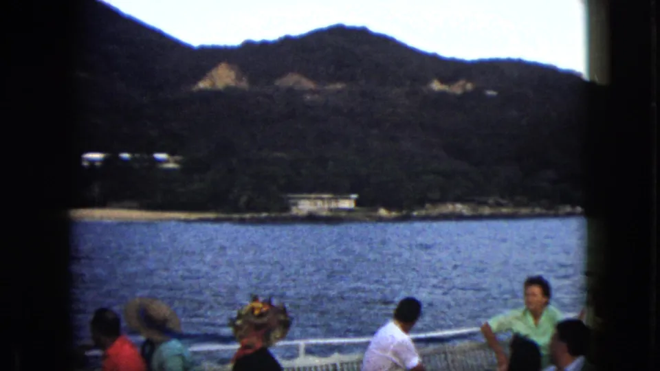 1962 Acapulco home-movie view from a boat, with passengers in hats before blue water and a distant coastal building.
