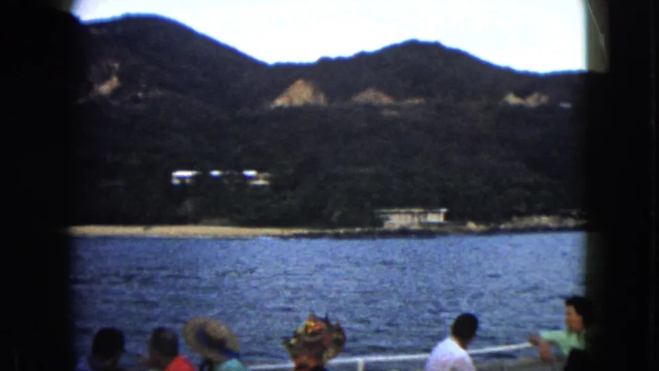1962 Acapulco frame showing tourists aboard a boat, blue water, dark green mountains, and small shoreline buildings.