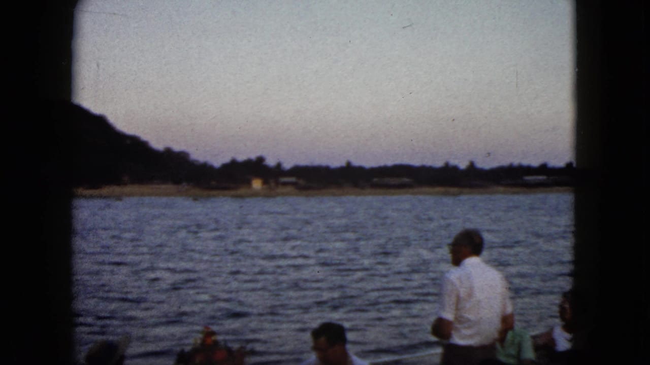 1962 Acapulco home-movie frame showing adults on a boat, including a man in a white shirt, with water and hillside shoreline at dusk.