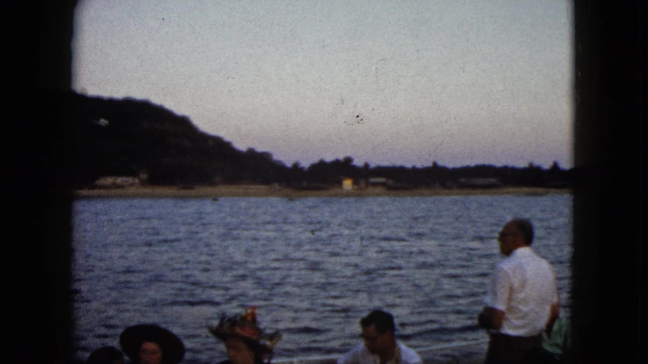 1962 Acapulco frame of adults aboard a boat, some wearing hats, with open water and a hillside shoreline in fading light.