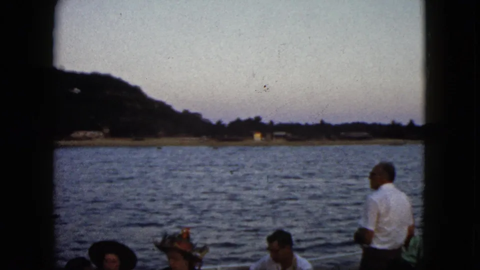 1962 Acapulco frame of adults aboard a boat, some wearing hats, with open water and a hillside shoreline in fading light.