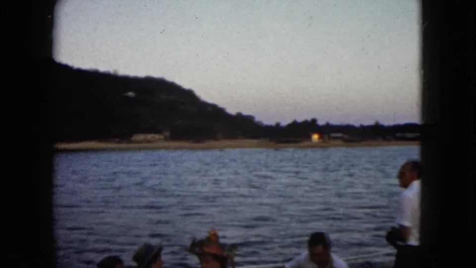 1962 Acapulco boat footage frame with partial adult passengers below, rippled water across the center, and a dark hillside at dusk.