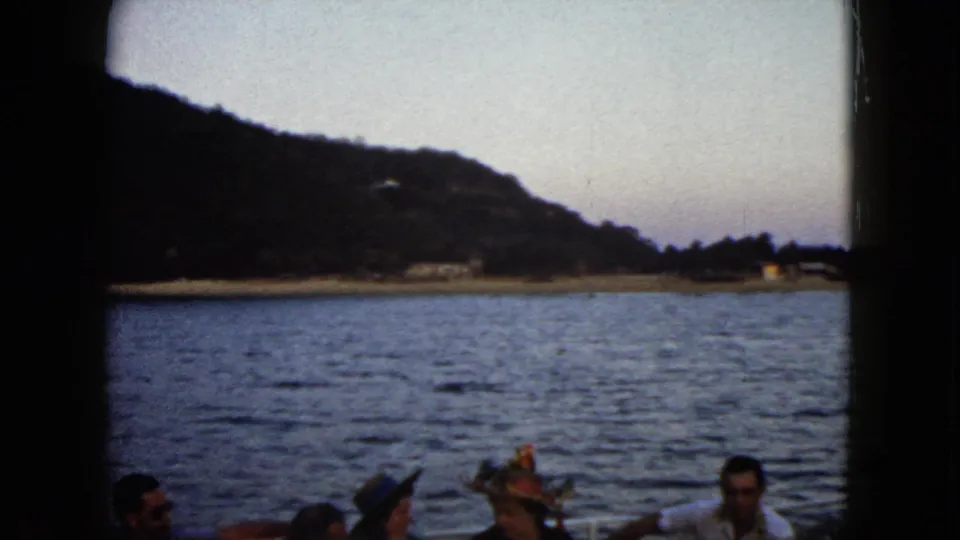 1962 Acapulco home-movie frame showing seated adults and hats on a boat against open water, shoreline buildings, and a dusky hillside.