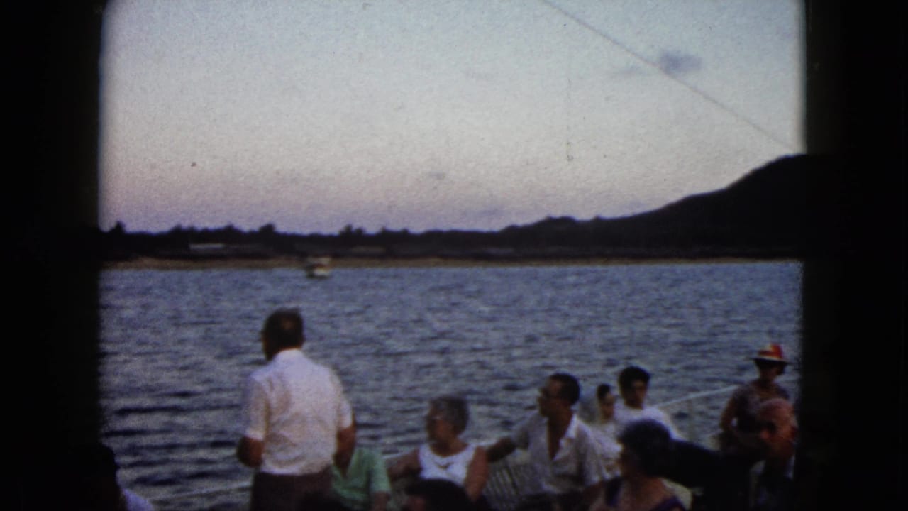 1962 Acapulco home-movie frame showing passengers on a tour boat looking toward water, shoreline, and hills in fading light.